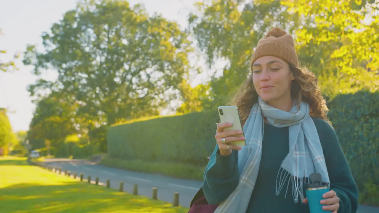 Smiling Young Woman Wearing Hat And Scarf Walking In Autumn Countryside Looking At Mobile Phone