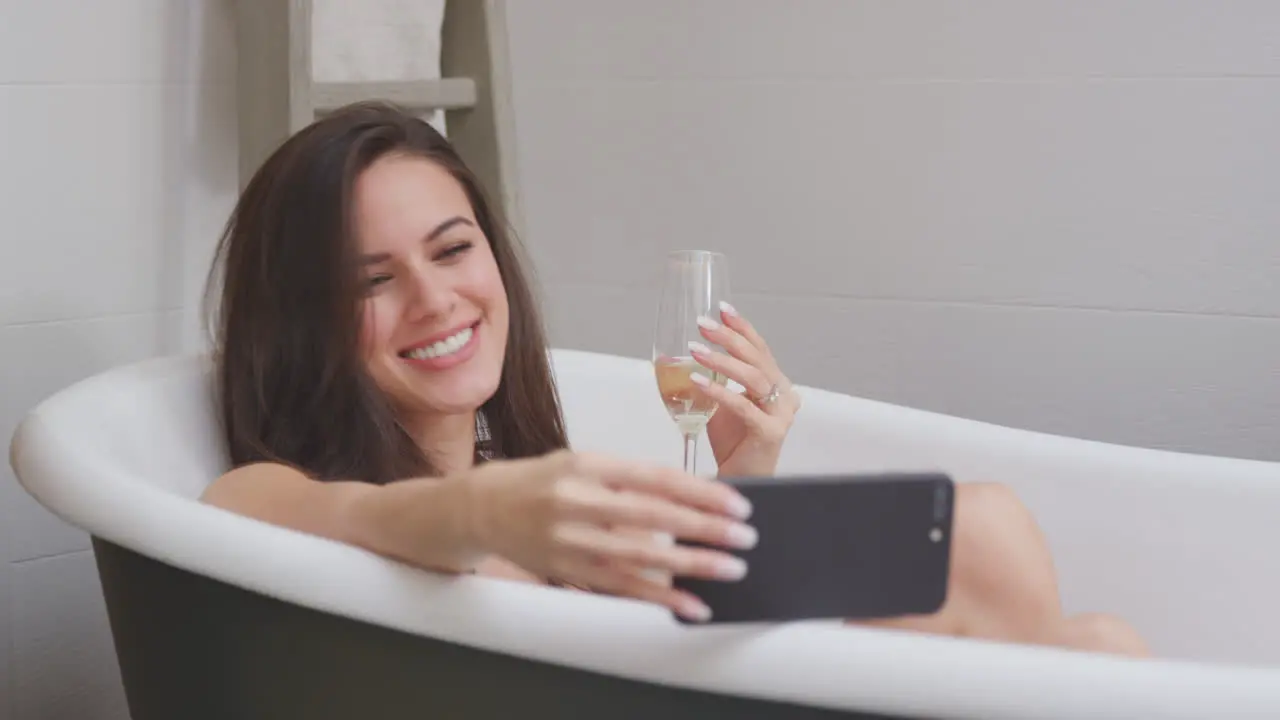 Woman Lying And Relaxing In Bath At Home Streaming Movie On Mobile Phone And Drinking Glass Of Wine