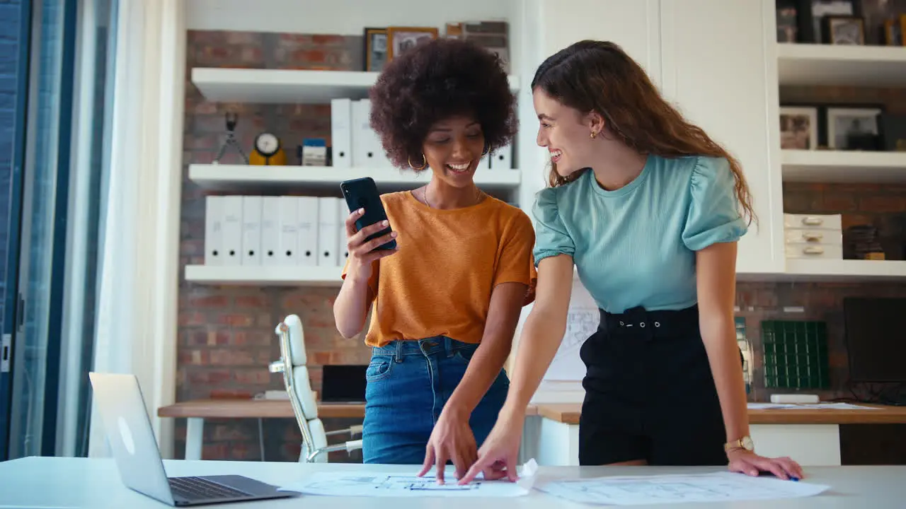 Two Female Architects With Laptop And Mobile Phone Meeting And Discussing Building Plans In Modern Office