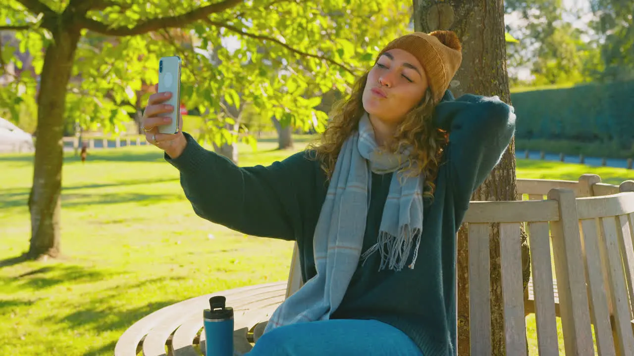 Woman Sitting On Bench In Autumn Park Posing For Selfie On Mobile Phone To Post To Social Media