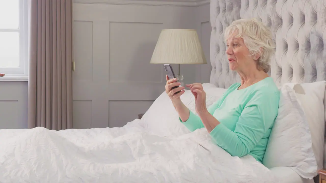 Smiling Senior Woman At Home Wearing Pyjamas In Bed Using Mobile Phone