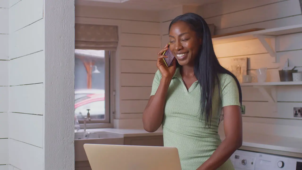 Young Woman Standing At Kitchen Counter With Laptop Working From Home Answering Call On Mobile Phone