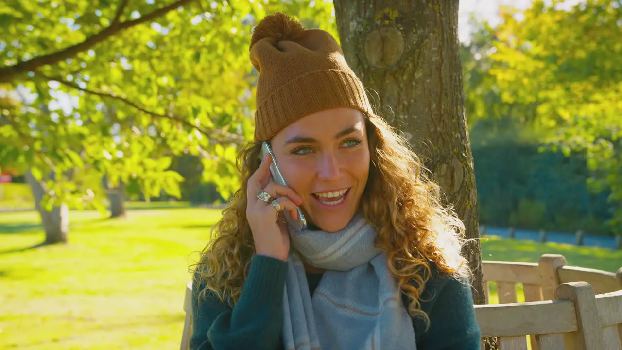 Smiling Young Woman Wearing Hat And Scarf Sitting On Bench In Autumn Park Talking On Mobile Phone