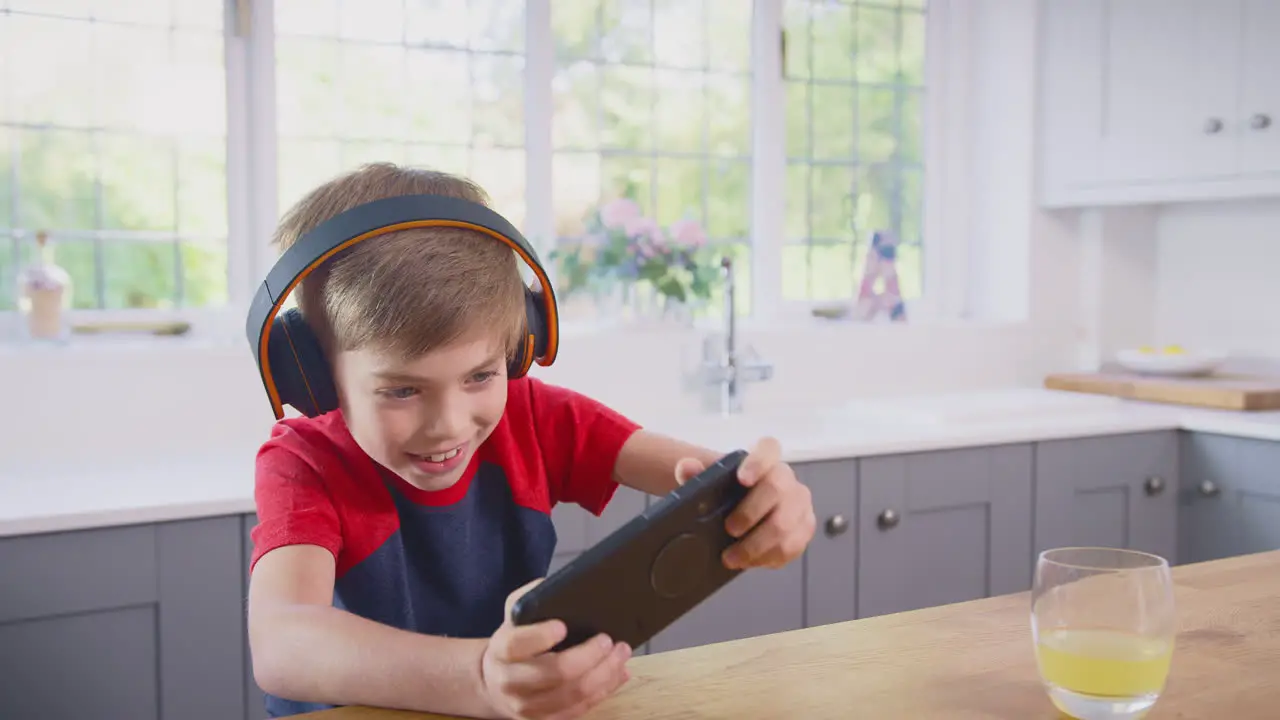 Boy At Home In Kitchen Playing Game On Mobile Phone Wearing Wireless Headphones