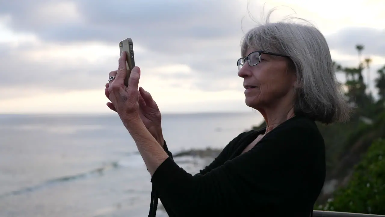 An old lady on vacation happy and smiling while taking a selfie with her smart phone on the ocean in Laguna Beach California SLOW MOTION