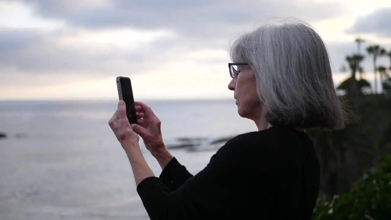 An old aging woman on vacation using her phone to take a picture at the beach over the ocean at sunset SLOW MOTION