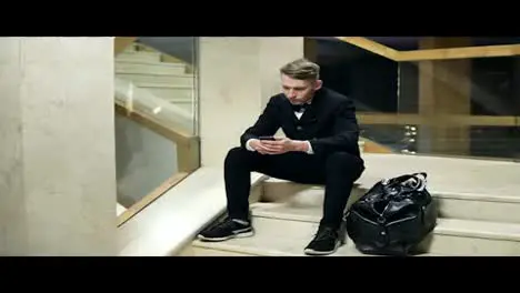 Young blonde man in black suit with big black leather bag using his smartphone and sitting on stairs in hotel Dialing a number