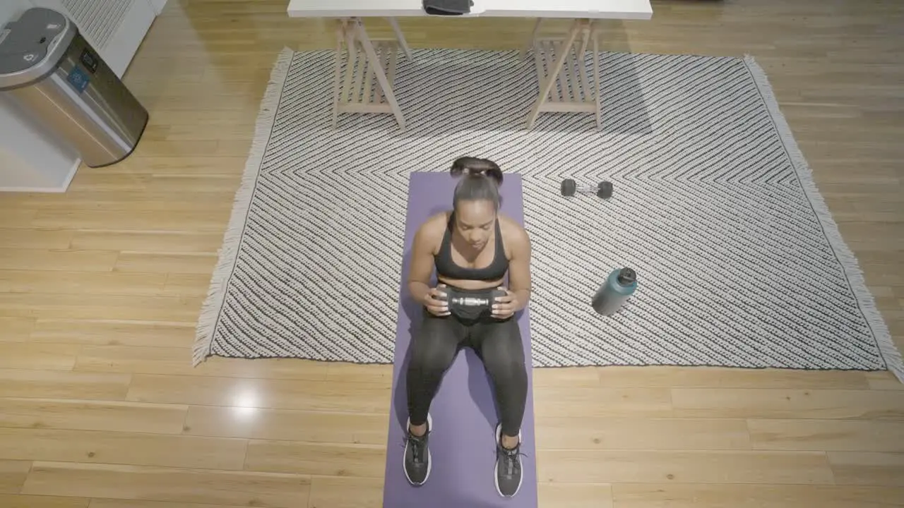 An overhead shot of a black female doing sit ups with weights in her hand on the floor of her kitchen