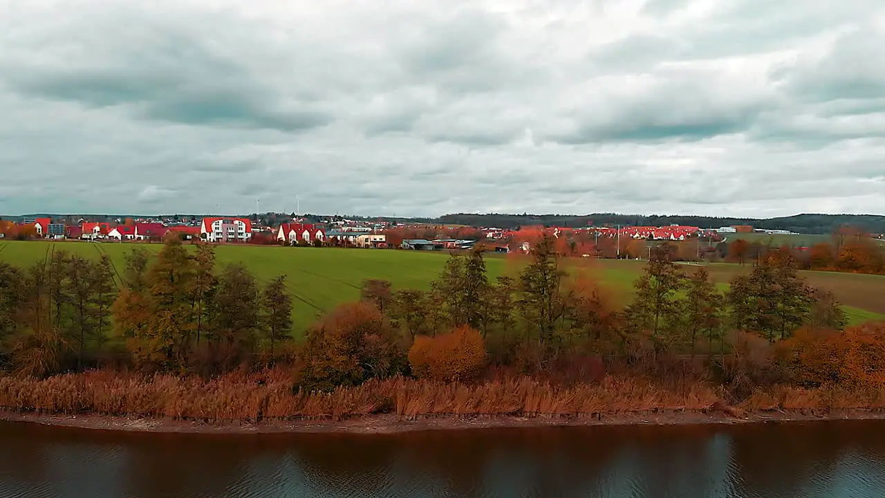 drone flies over the shore of a lake there is a green meadow a small town and a cloudy blue sky