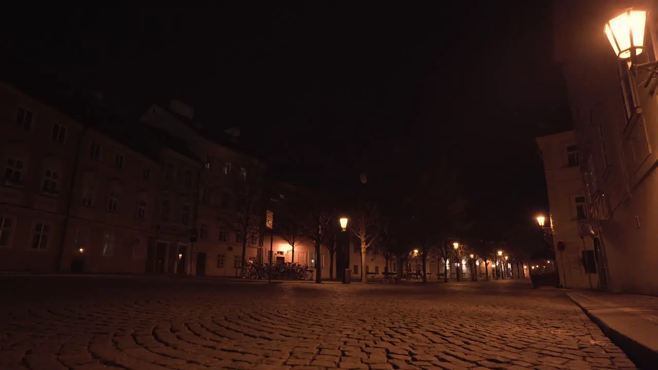 Empty city square Kampa of paving stones in Prague Czechia with trees old houses and street lanterns shining above during a Covid-19 lockdown at night illuminated by streetlights slow pan shot