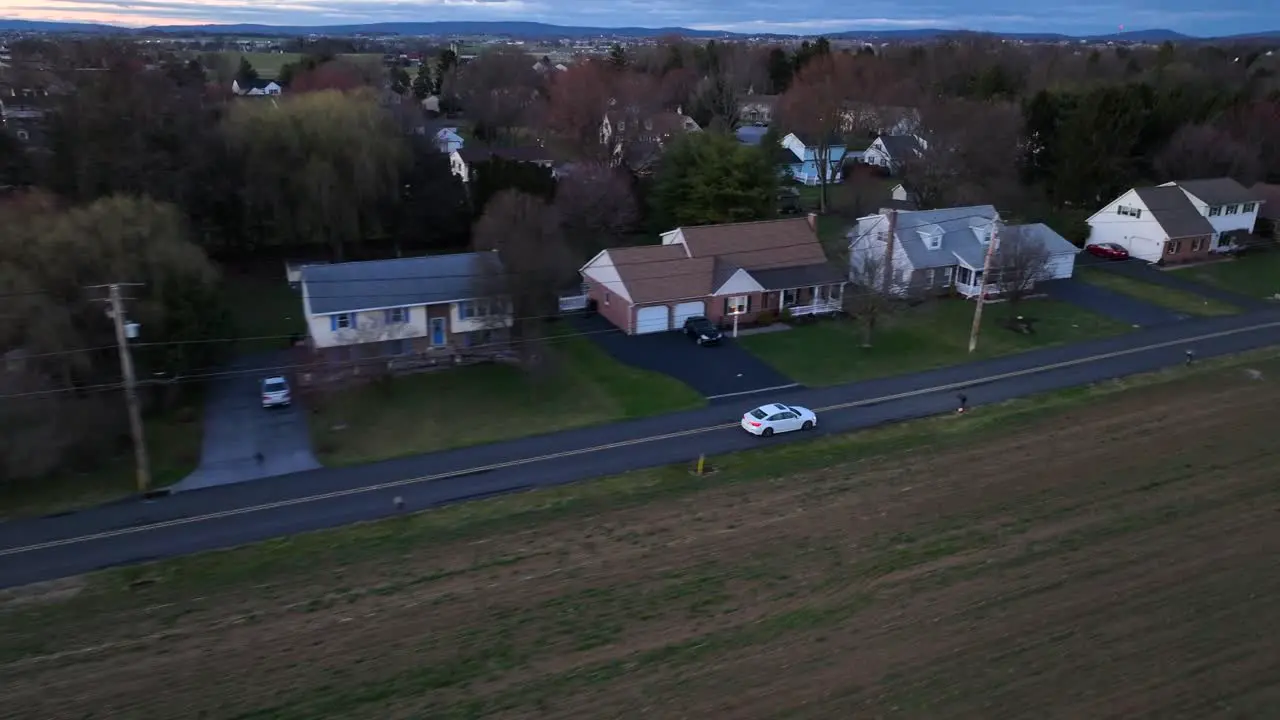 High speed tracking aerial shot of white sedan driving on rural street next to neighborhood at night