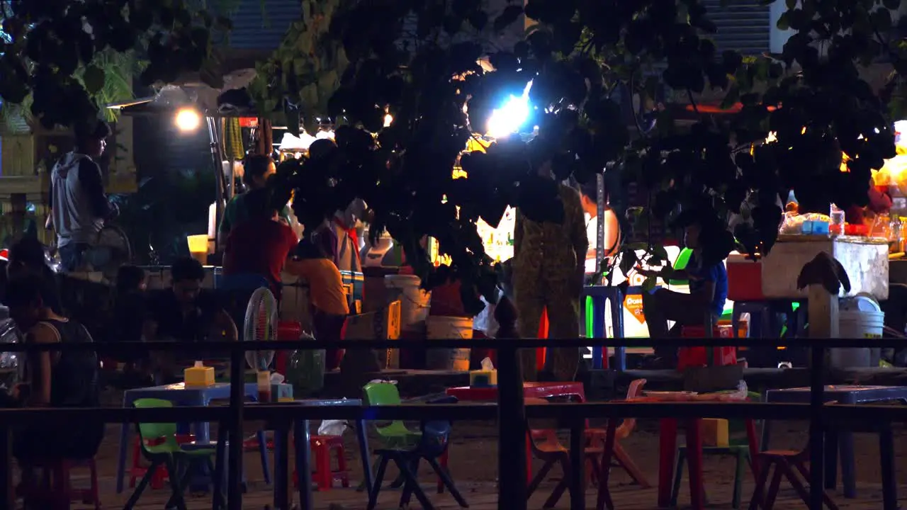 Wide Shot of Small out Side Eating Place with Plastic Chairs and Tables with People Walking Around One swinging in a Hammock at night time with traffic in the background