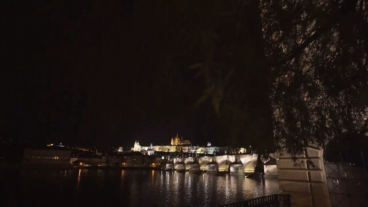 The Prague Castle and Charles bridge over river Vltava in the historical centre of Prague Czechia lit by lights at night shot from the other side of the river slowly tilting through willow branches