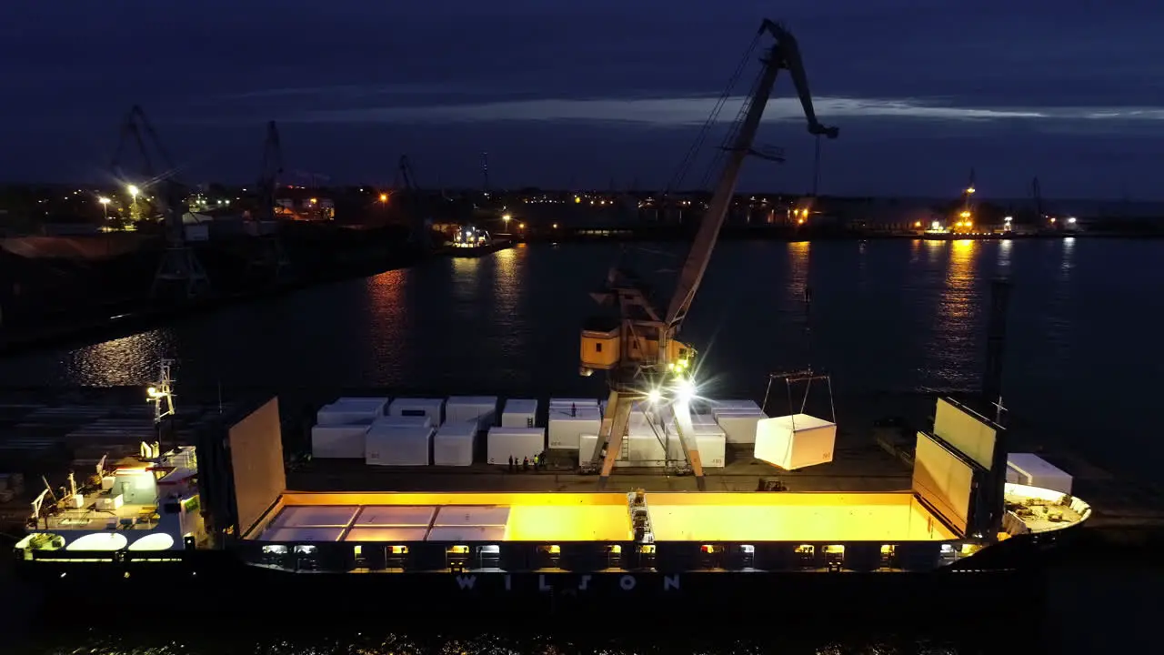 Shipping containers being loaded on a cargo vessel at night with a crane ascending aerial view