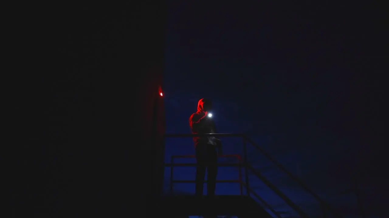 Mysterious hooded man on top of metal stairs shining a flashlight around at night time