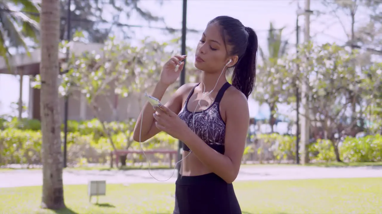 Cheerful Asian woman in earphones browsing smartphone in hand in park