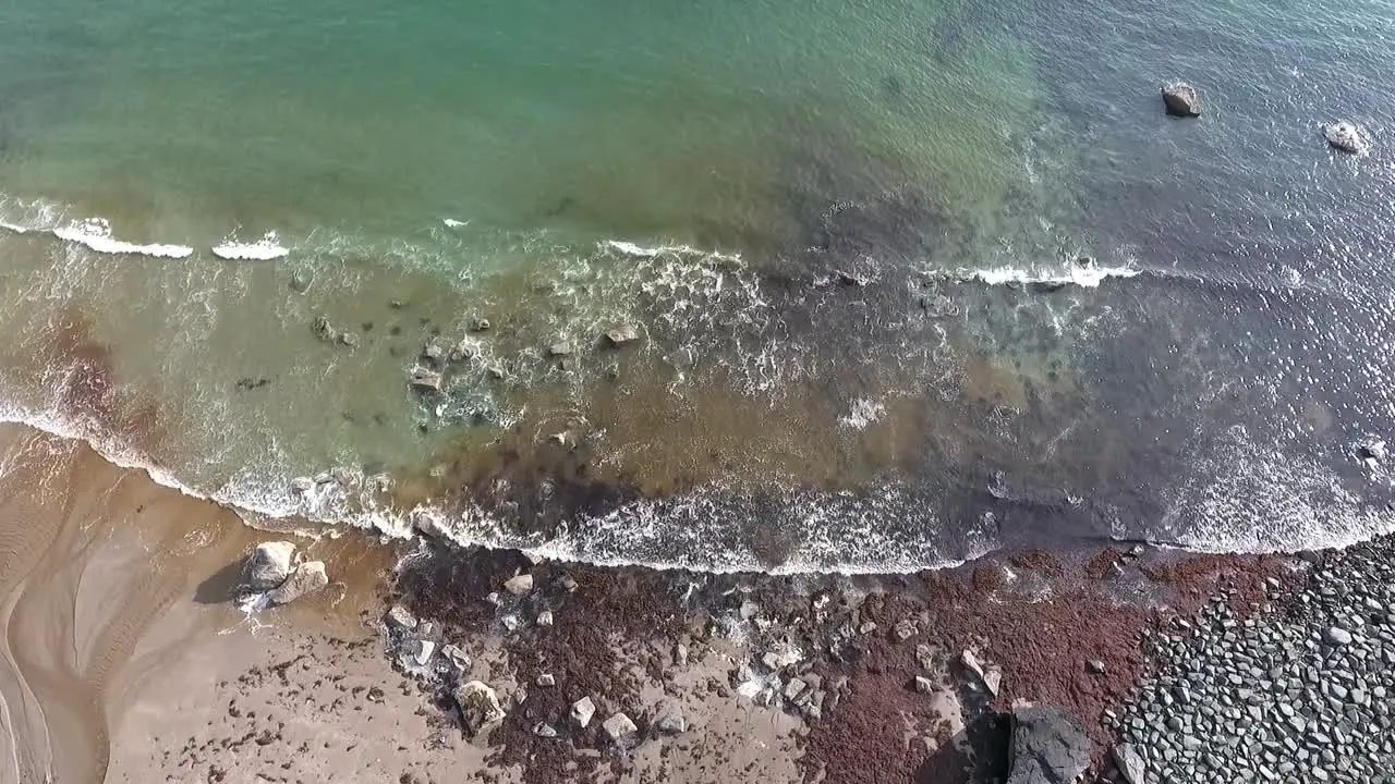 Aerial overhead descending view of green sea and white foaming waves gently breaking on the rocky sandy shore in summer