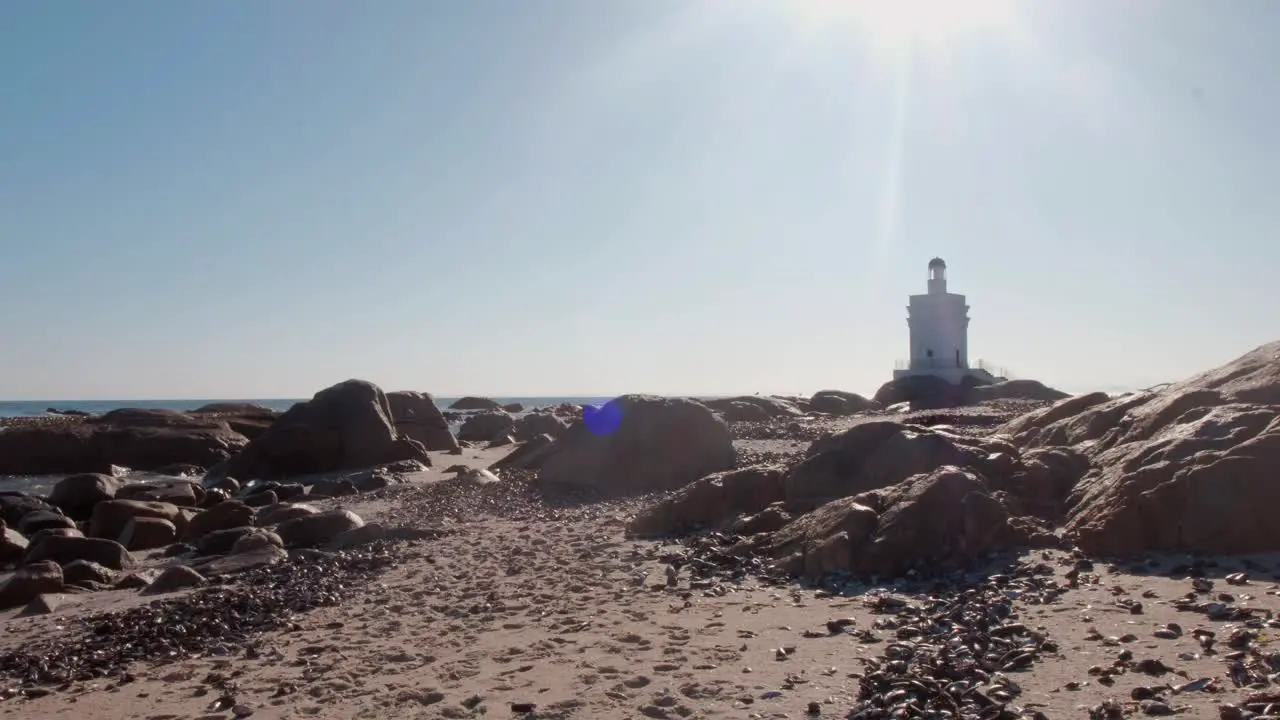 View of a lighthouse on the beach with waves softly crashing against the rocks in the midday sun