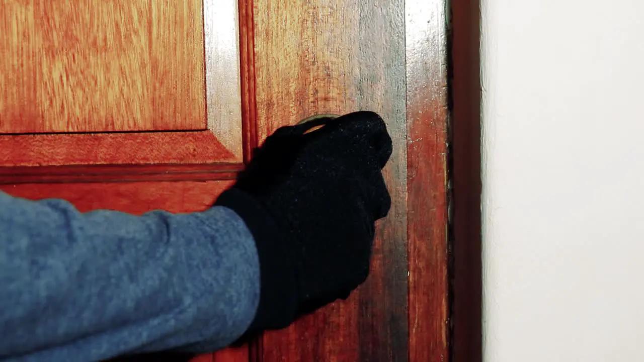 Man with a black glove on slowly closes a brass door knob on a wooden door