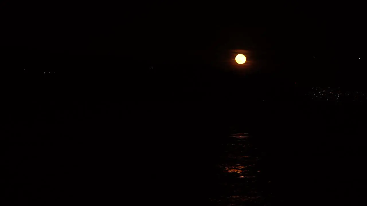 Full moon reflection in small waves on water surrounded by deep black background small city lights in the distance