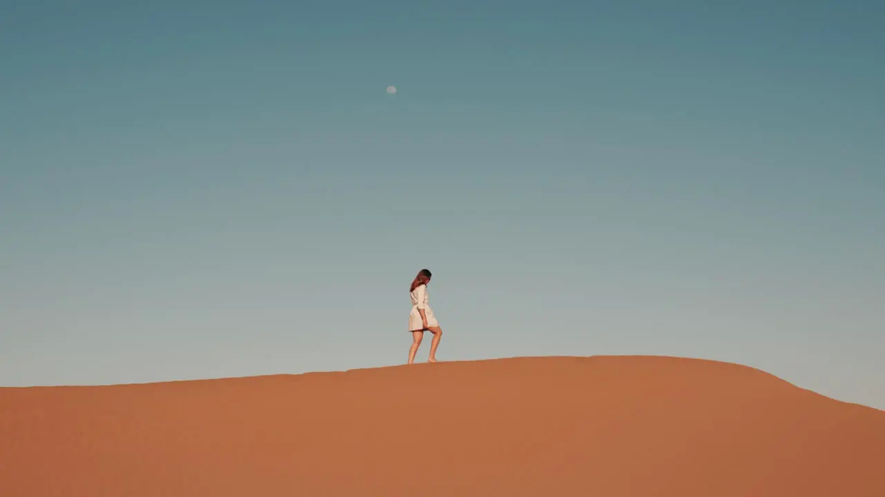 Young caucasian woman walking on top of a desert dune during sunset with moon in the background in Zagora Morocco