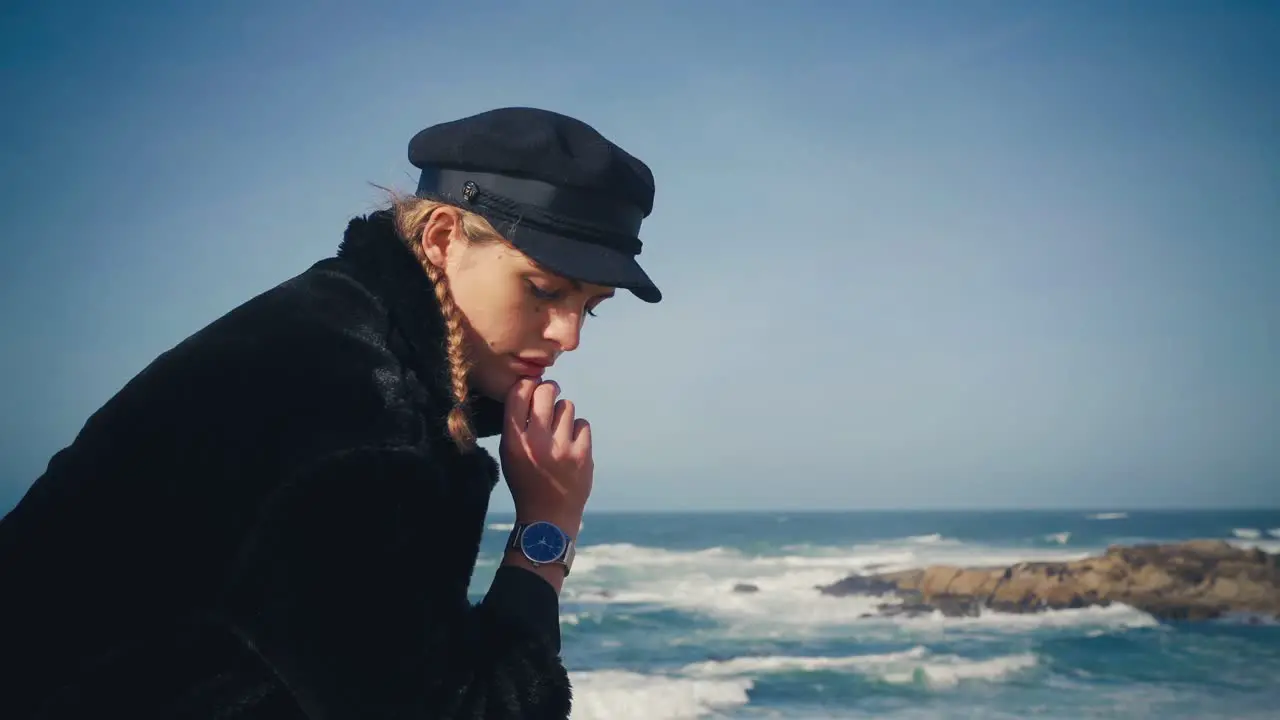 4K UHD Cinemagraph of a young brunette woman sitting by the sea with a black coat and black hat with the waves rushing in the background