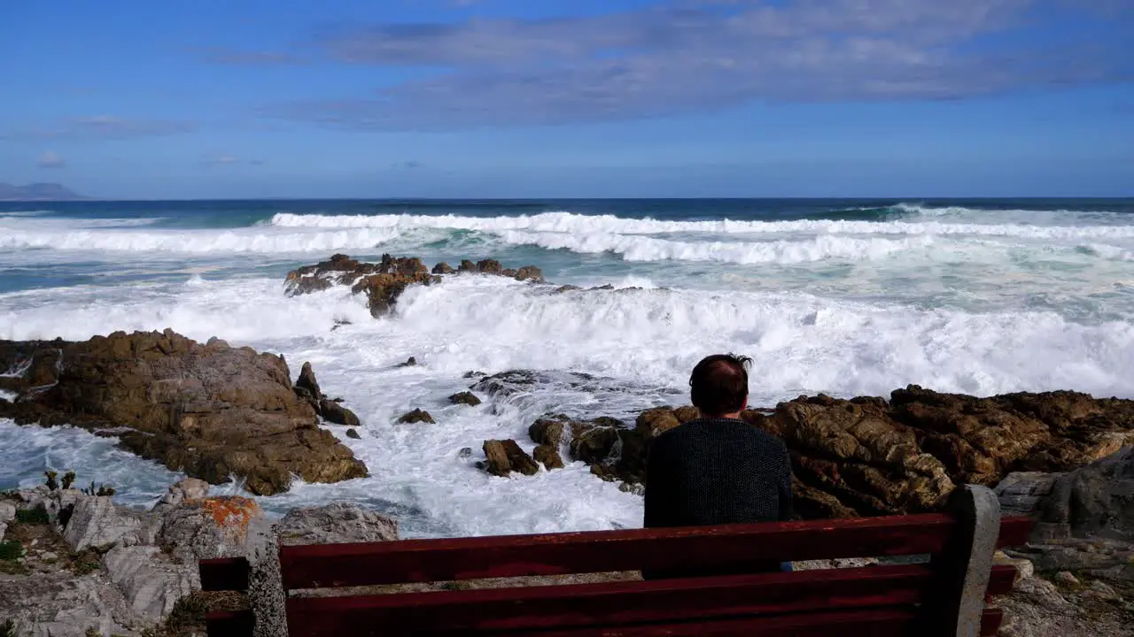 Pensive man on bench overlooking ocean and waves breaking onto rocky coastline