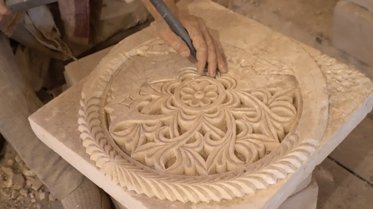 we see a stone ornament artist working on a white stone with a hammer in one hand and a nail in the other