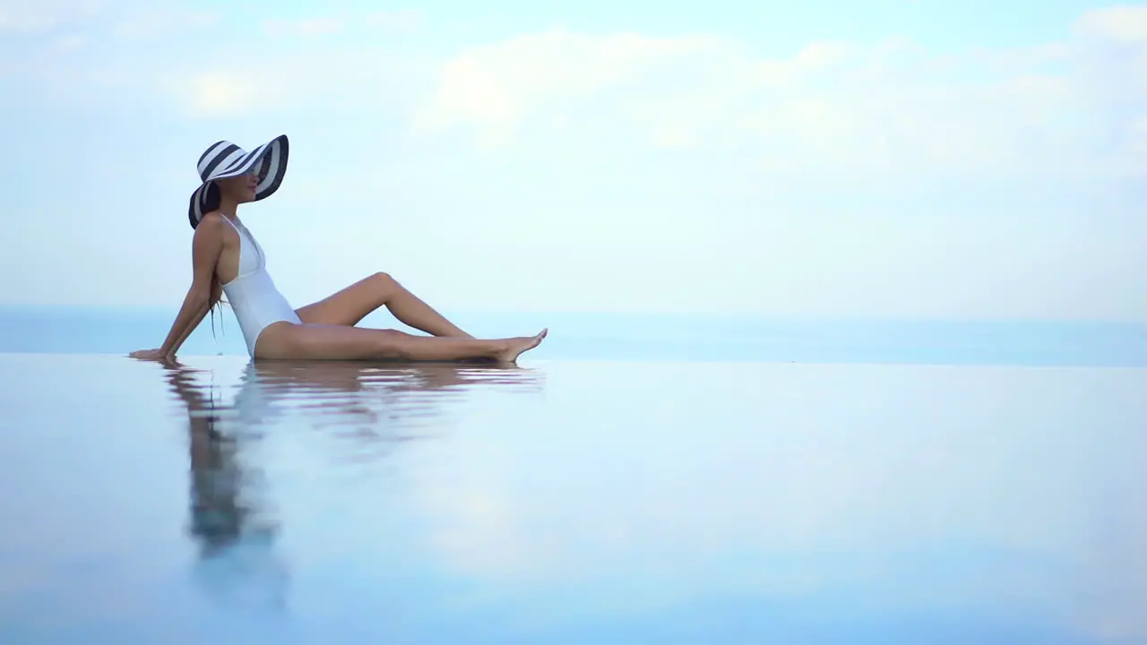 Petite Woman in Swimsuit and Summer Hat Sitting on Infinity Pool Collided With Sea Skyline