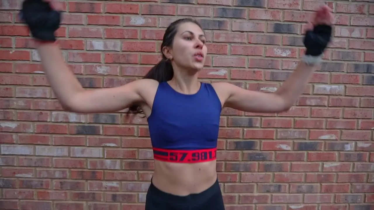 Young Hispanic brunette adult female in sportswear doing jumping jacks routine in an outside urban scene in front of a brick wall