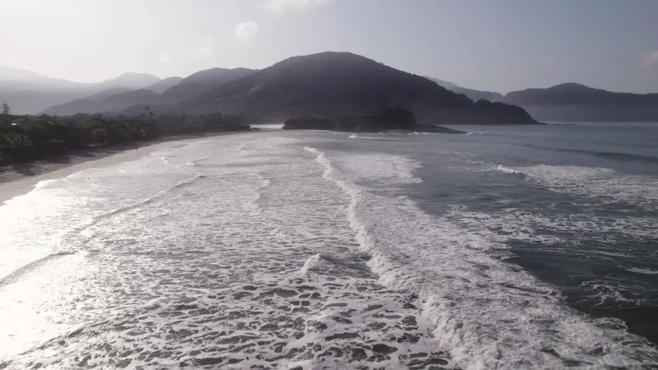 Drone flying back at the shore showing the waves sand and the atlantic rainforest at the backgound with the light of the morning and fog