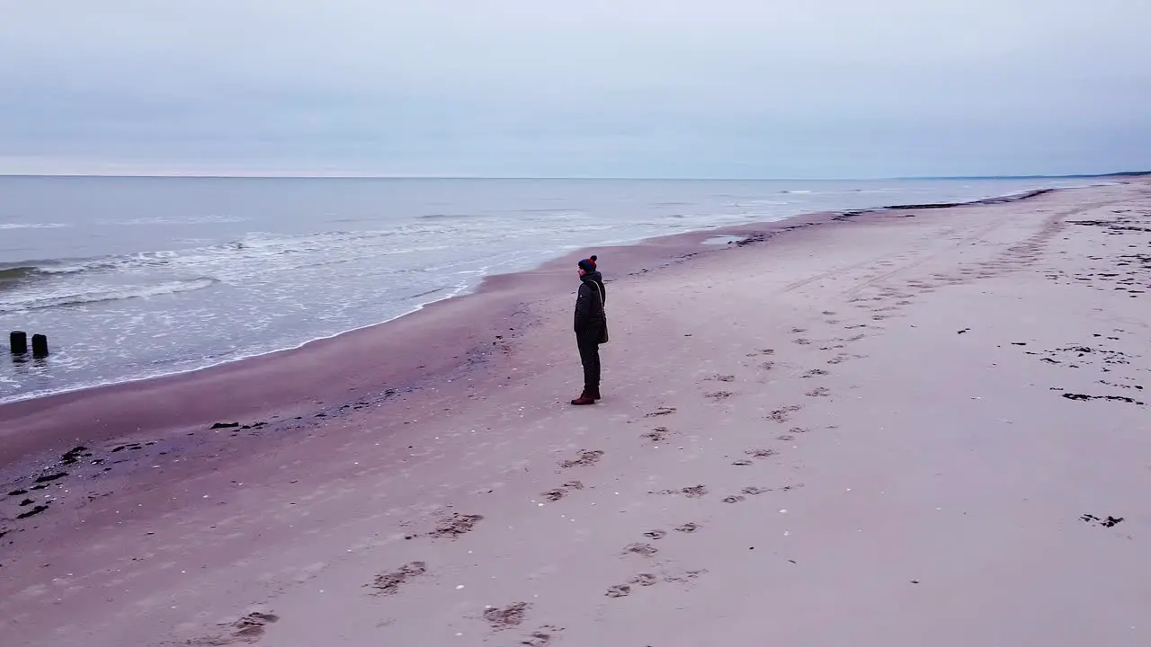 Beautiful aerial view of a caucasian man standing on the white sand beach old wooden pier at the Baltic sea coastline overcast day coastal erosion calm seashore wide angle drone shot orbit right