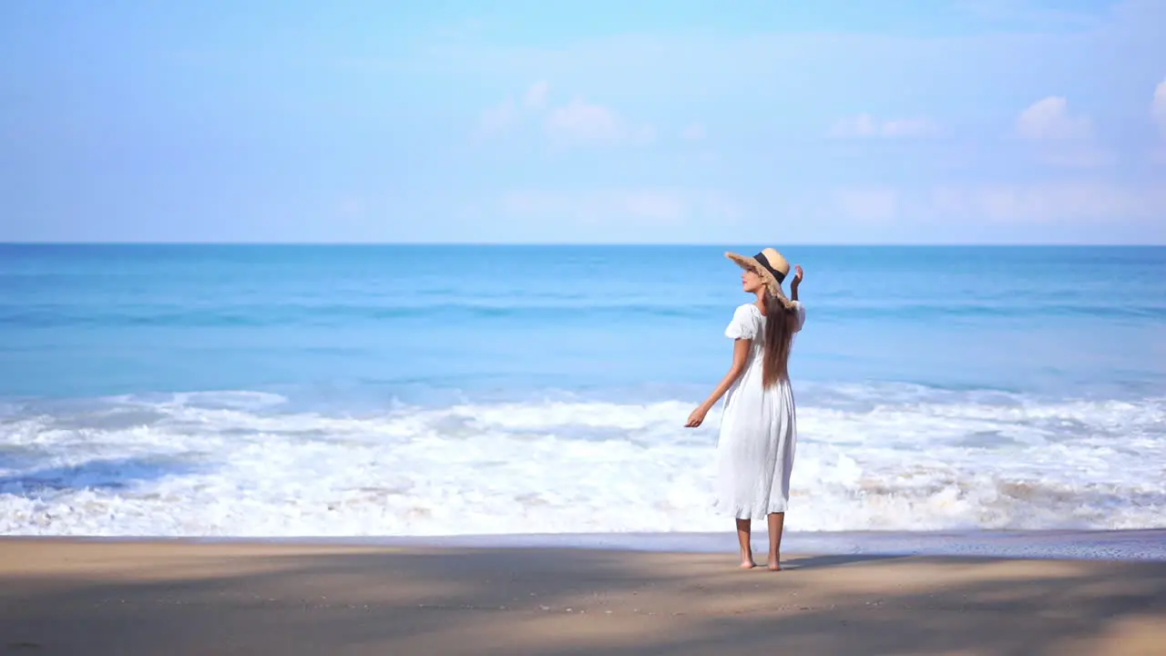 Asian Woman standing on the beach by the sea water when big foamy tides roll on sandy beach in summer wearing a white sundress and straw hat slow-motion static shot