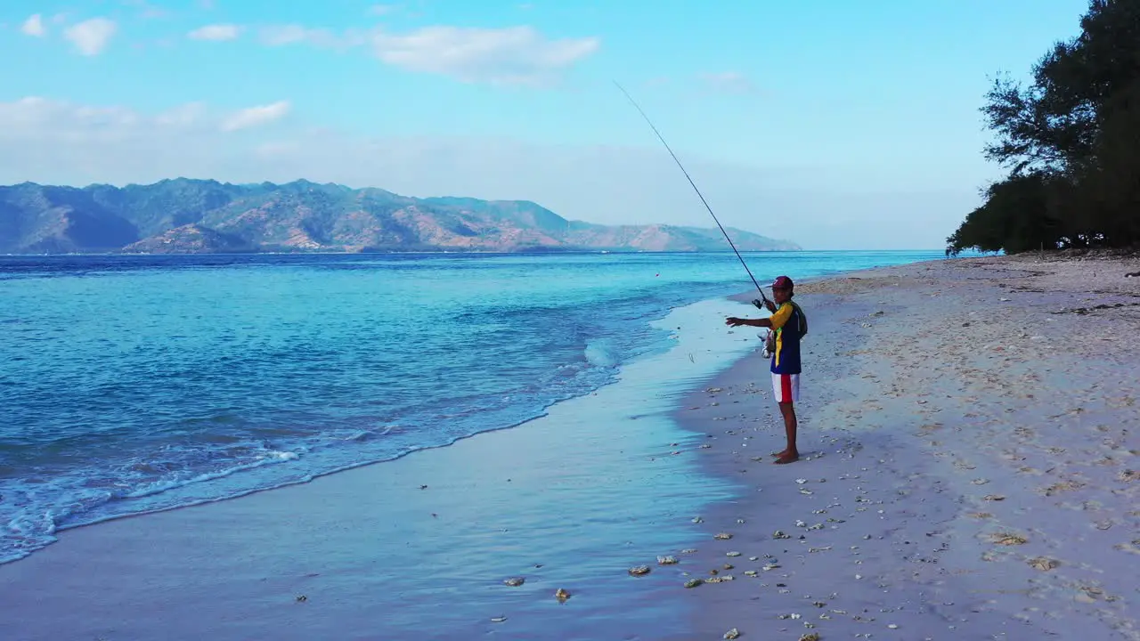 Young man fishing a small fish rolling rod line from blue sea on empty beach at sunset low light Bali