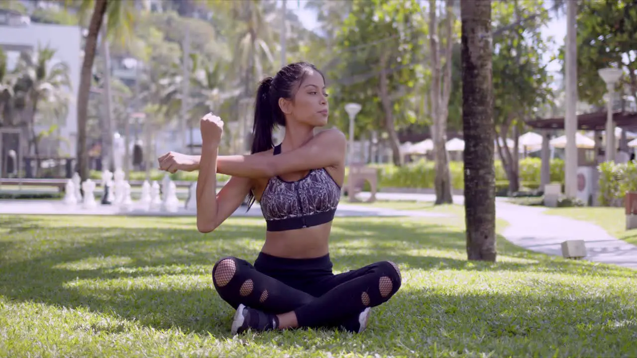 Sporty woman sitting with crossed legs and stretching arms in park