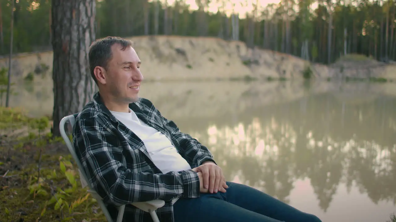 relaxed adult man is sitting in armchair on shore of calm picturesque lake in forest and admiring nature and calmness