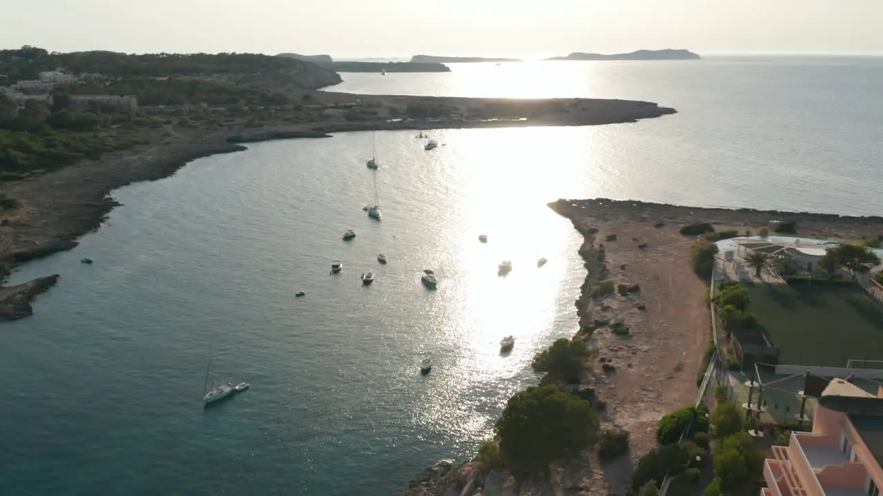 Aerial view of boats parked in a silent and calm sea with empty resort at shore with a football ground and greenery in Ibiza in Spain