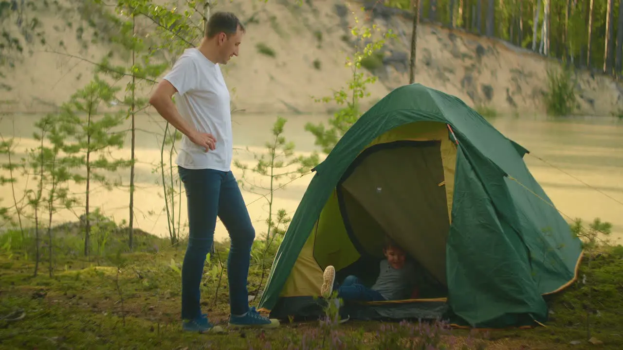 Boy climbs into a tent on the shore of the lake while traveling and relaxing with his father in nature