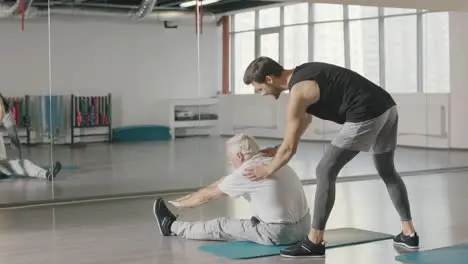 Pensioner man doing stretching exercise with fitness instructor in gym together