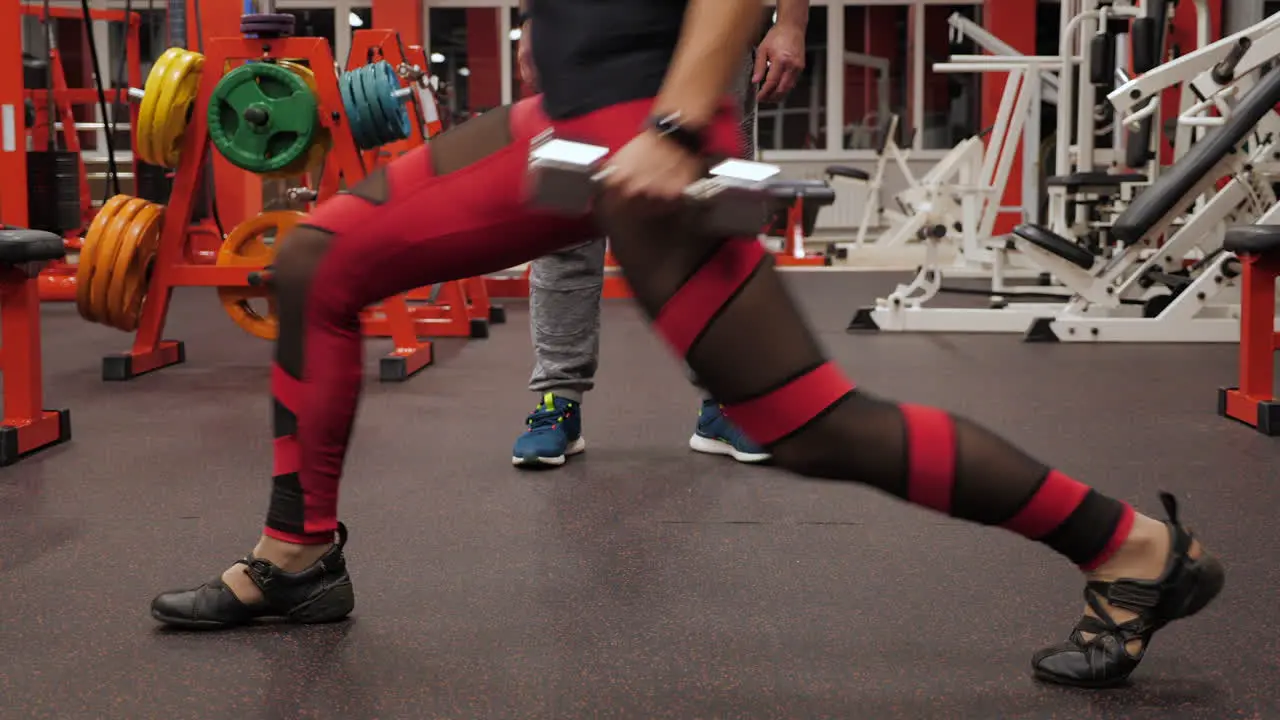 A woman doing leg lunge with weights in a gym under the supervision of a coach