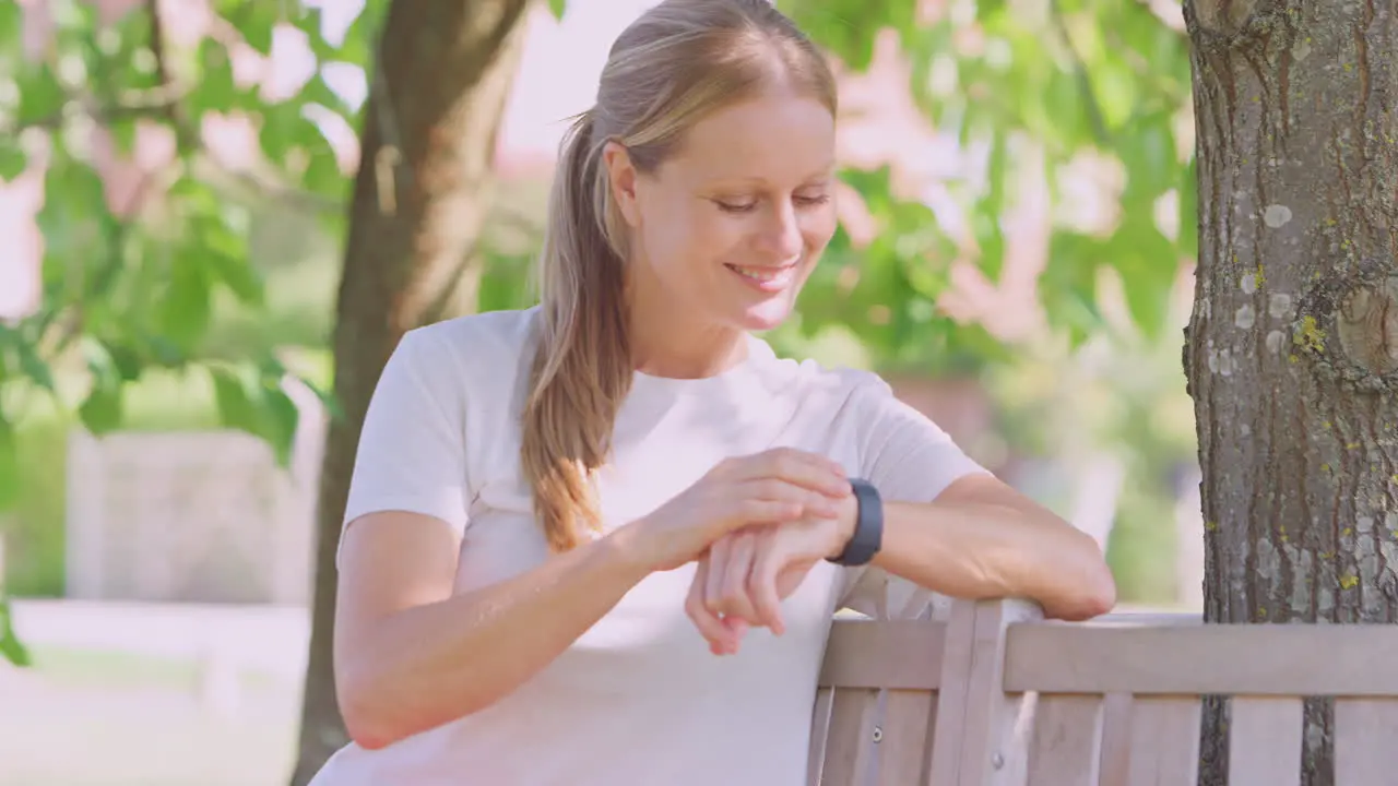 Woman Wearing Fitness Clothing Sitting On Seat Under Tree Checking Activity Monitor On Smartwatch