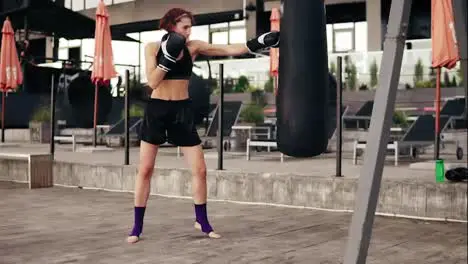 Strong athletic female boxer in gloves exercising with a bag She hits a bag with her leg Workout outside Female boxer