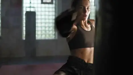 Strong athletic female kickboxer hits a boxing bag with her leg She is exercising with a boxing bag in dark gym with smoke