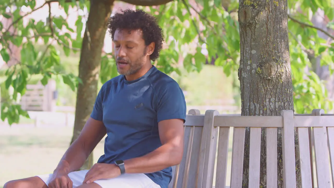 Mature Man Wearing Fitness Clothing Sits On Seat Under Tree Checking Activity Monitor On Smartwatch