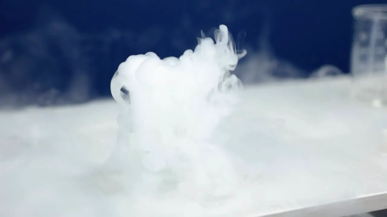 A beaker in a laboratory emits a cascade of white smoke bubbling and swirling mysteriously against a blue background