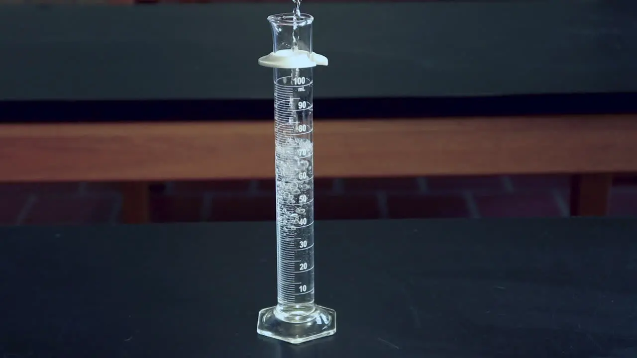 Clear liquid being poured into a 100mL graduated cylinder on a black lab bench in a high school chimistry classroom