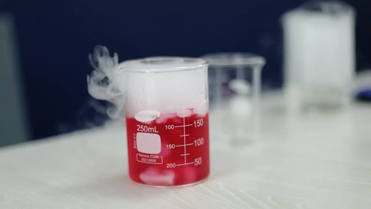 In striking slow-motion a glass beaker filled with a luminous red liquid undergoes a chemical transformation as tendrils of white smoke rise embodying the intricacy of scientific wonders