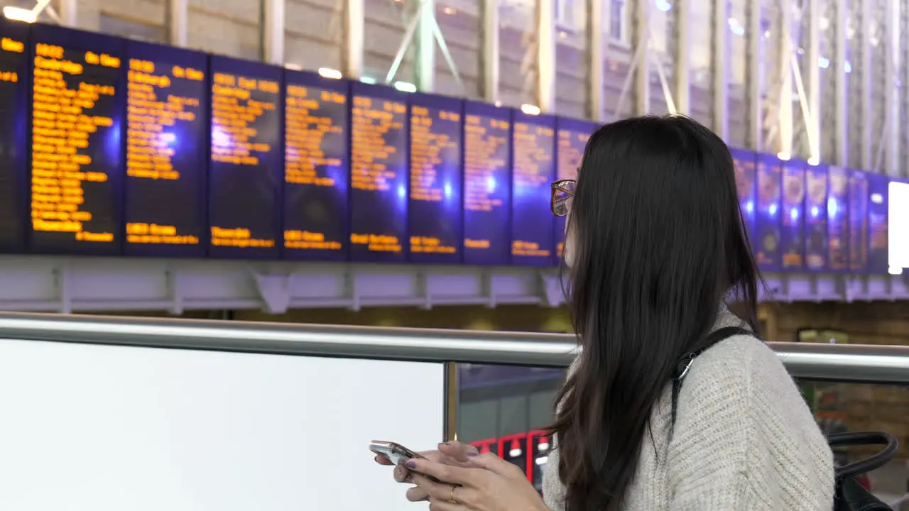 Young woman on her phone checks the train station departure board before leaving to catch her train