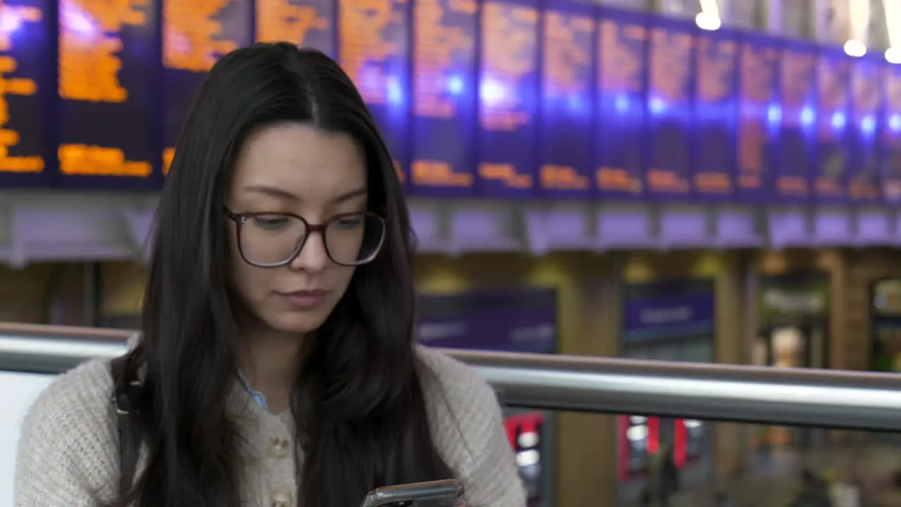 Young woman looking at her phone while waiting for her train station departure boards behind her