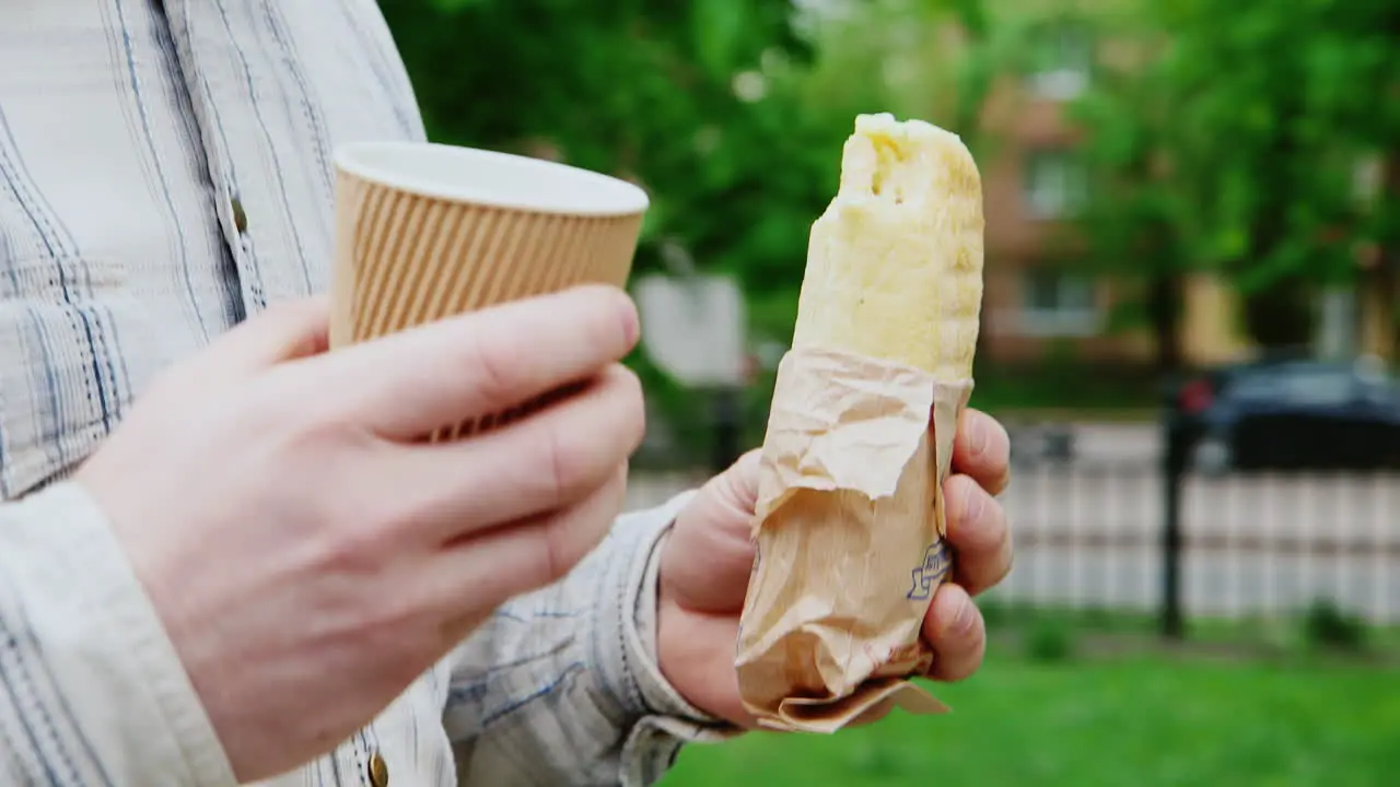 A Man Walks Down The Street With A Coffee And A Sandwich In The Frame Only Hands Visible